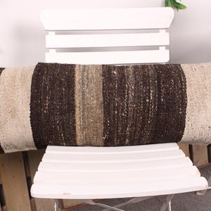 May include: A long, rectangular pillow with a woven texture. The pillow features alternating stripes of dark brown, beige, and light brown. It is placed on a white chair, suggesting a decorative or functional home decor item.