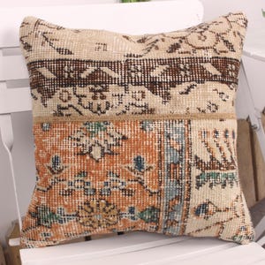 Puede incluir: Cojín decorativo cuadrado con diseño de patchwork. La sección superior presenta un patrón beige y marrón, mientras que la sección inferior muestra un diseño floral naranja y turquesa. El cojín está colocado sobre una silla blanca.