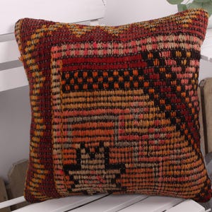 May include: Square decorative pillow with a woven, textured design. The pillow features a mix of colors, including red, orange, and black, creating a geometric pattern. The pillow is placed on a white surface.