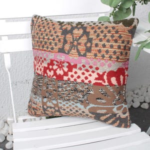 May include: A colorful patchwork throw pillow with a geometric pattern in shades of red, pink, brown, and gray. The pillow is sitting on a white chair.