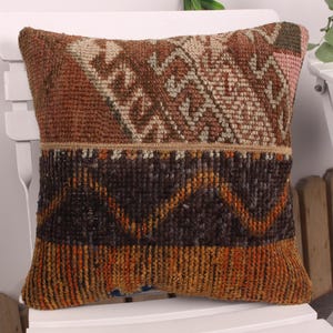 May include: Square decorative pillow with a woven, textured design. The pillow features a patchwork of colors including brown, orange, and black. The top section has a geometric pattern, while the bottom has a zig-zag design.