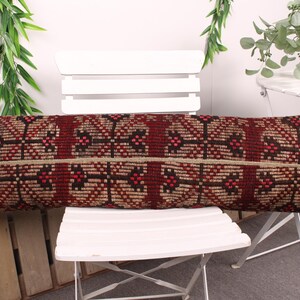 May include: A long, rectangular pillow with a geometric pattern in shades of burgundy, brown, and beige. The pillow is placed on a white chair. The pillow's dimensions are approximately 36 inches long.