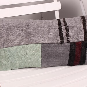 May include: A rectangular decorative pillow with a patchwork design. The pillow features sections of gray, light green, and dark gray fabric. Dark brown and red stripes accent the gray sections. The pillow is placed on a white chair.