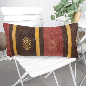 May include: A long, rectangular pillow with a brown, red, and yellow striped pattern. The pillow has a geometric design in the center of each stripe. The pillow is sitting on a white chair.