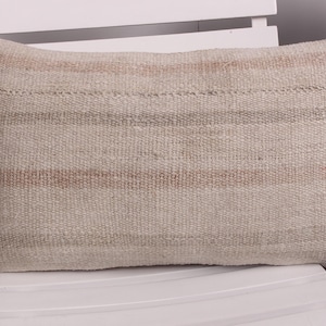 May include: Rectangular beige pillow with horizontal stripes in shades of brown and tan. The pillow has a woven texture and is placed on a white chair. The pillow is a decorative item for a bed or sofa.
