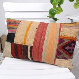 May include: Rectangular patchwork pillow with a variety of colors including beige, red, orange, and purple. The pillow is made of woven fabric and has a geometric pattern on one end. It is placed on a white chair.