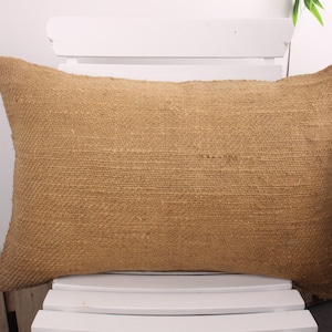 May include: A rectangular, woven pillow in a natural tan color. The pillow has a textured surface and is placed on a white chair. The pillow is a decorative item for a home.