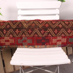 May include: A rectangular decorative pillow with a patterned design. The pillow features a combination of brown, red, and gold colors. The pillow is placed on a white chair.