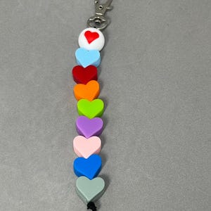 Peut inclure: Un porte-clés coloré avec des perles en forme de cœur de différentes couleurs : rouge, bleu, orange, vert, violet et rose. Il est doté d'un fermoir argenté en haut et d'une ficelle noire en bas. Les perles sont en silicone.