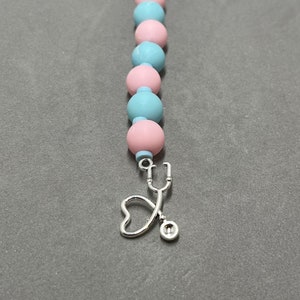 Puede incluir: Un llavero de cuentas rosa y azul con un dije de estetoscopio plateado y un corazón.