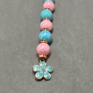 May include: A pink and blue beaded charm with a blue flower charm at the end. The beads are iridescent and have a metallic sheen.