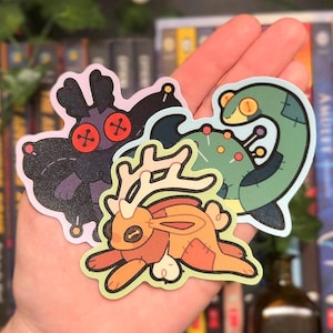 May include: Three die-cut vinyl stickers featuring stylized creatures. One is a dark purple moth with red button eyes, one is a green sea serpent with yellow button eyes and pins, and one is an orange jackalope with a button eye and stitched patches.