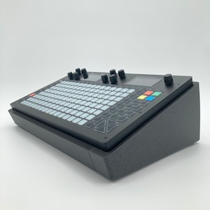 May include: A black music production controller with a grid of buttons and knobs. The controller is sitting on a black foam stand.