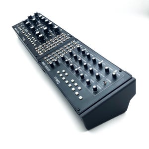 May include: A black Moog synthesizer with numerous knobs, buttons, and input jacks. The synthesizer has the Moog logo on the front and text indicating various functions. The device is rectangular and sits on a white surface.