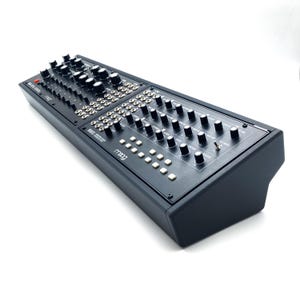 Puede incluir: Un sintetizador negro con numerosas perillas, botones e interruptores. El dispositivo tiene forma rectangular con un panel frontal ligeramente inclinado. La marca "moog" es visible en la parte delantera. El sintetizador está diseñado para crear y manipular sonidos electrónicos.