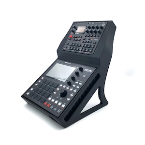 May include: Black Akai MPC One music production centre with a built-in screen, buttons, knobs, and pads. The device is mounted on a black stand. The Akai logo is visible on the front.