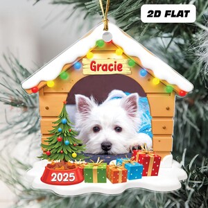 May include: A 2D flat Christmas ornament featuring a white dog named Gracie inside a dog house. The dog house is decorated with a string of colorful lights and a small Christmas tree. The ornament includes the year 2025 and several wrapped gifts.