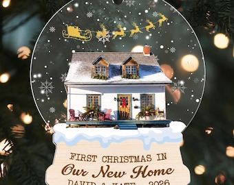 May include: A circular glass ornament featuring a white house with a snowy roof, a porch with red chairs, and a golden silhouette of Santa and reindeer. The base displays the text: FIRST CHRISTMAS IN Our New Home DAVID & KATE - 2026.