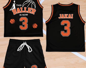 May include: Black basketball jersey and shorts set with orange trim. The jersey has the text "THIS BALLER IS NOW 3" and a basketball graphic. The shorts have the number 3. The back of the jersey has the name "JAKAI" and the number 3.