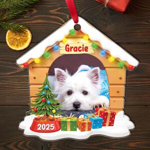 May include: A personalized dog house Christmas ornament featuring a white dog named Gracie. The ornament includes a miniature Christmas tree, presents, and a bowl with "2025" on it. The ornament is trimmed with snow and colorful lights.
