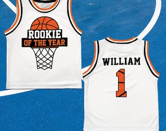 May include: White basketball jersey with orange and black trim. The front features a basketball graphic with the text "ROOKIE OF THE YEAR" above a basketball net. The back displays the name "WILLIAM" and the number "1" in orange.