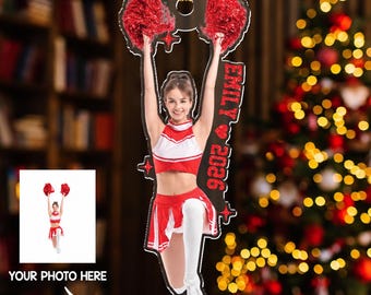 May include: A custom acrylic ornament featuring a cheerleader in a red and white uniform holding red pom-poms. The ornament includes the name EMILY, a heart icon, and the year 2026 printed in red text. It hangs from a gold string loop.