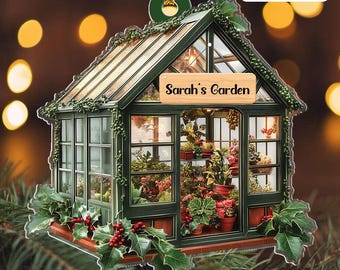 May include: A decorative greenhouse ornament with the text "Sarah's Garden." The green structure features multiple windows, a glass roof, and is filled with potted plants. Holly and berries adorn the base, with a gold hanging loop at the top.