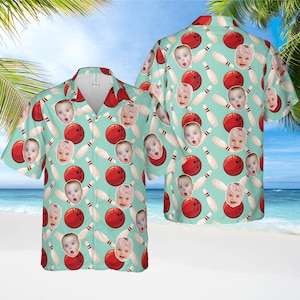 May include: A short-sleeved button-down shirt with a bowling theme. The shirt features a light teal background with a pattern of red bowling balls, white bowling pins, and baby faces. The shirt has a classic collar and a relaxed fit.