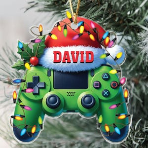 May include: A green video game controller Christmas ornament with the name "DAVID" on a Santa hat. The ornament is decorated with colorful Christmas lights and holly.
