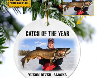 May include: White ceramic ornament with a photo of a person holding a large fish. Text on the ornament reads "CATCH OF THE YEAR", "YUKON RIVER, ALASKA 2025". Includes a gold string for hanging. A smaller image of the person and fish is in the upper right corner.