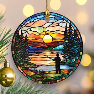 May include: Round Christmas ornament with a stained glass design. The ornament features a sunset scene with a fisherman, trees, and a lake. The ornament is trimmed in black and has a gold hanging cord.
