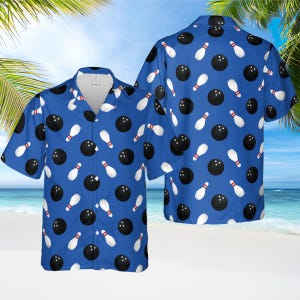 May include: Blue short-sleeved button-down shirt with a bowling theme. The shirt features a repeating pattern of black bowling balls and white bowling pins with red accents. The shirt is displayed on a beach background.