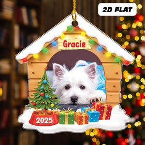 May include: A 2D flat Christmas ornament shaped like a dog house, featuring a white dog named Gracie. The ornament includes a Christmas tree, presents, and a dog bowl with "2025" on it. The ornament is decorated with colorful lights.