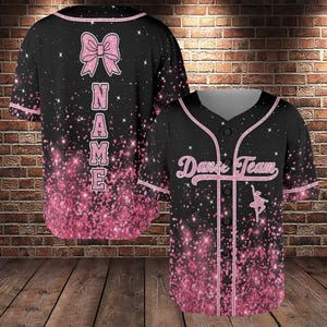 May include: Black baseball jersey with pink accents and a glittery design. The front features "Dance Team" and a ballerina silhouette. The back has a pink bow and the word "NAME" in pink letters. The jersey has a V-neck and button-up front.