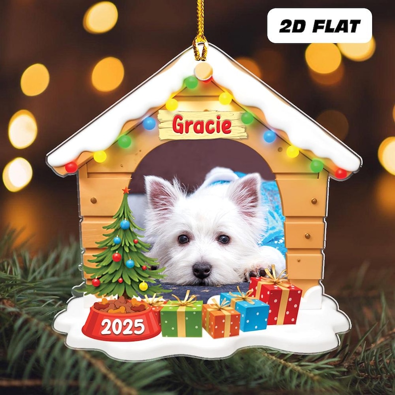 May include: A dog house-shaped Christmas ornament featuring a white dog named Gracie. The ornament includes a small Christmas tree, a food bowl with "2025", and wrapped gifts. The ornament is decorated with colorful lights.