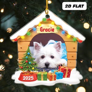 May include: A personalized Christmas ornament shaped like a dog house, featuring a white dog named Gracie. The ornament includes a small Christmas tree, presents, and a bowl with "2025" on it. The ornament is decorated with colorful lights.