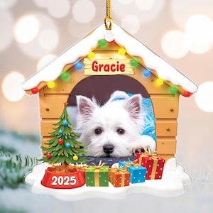 May include: A dog house Christmas ornament featuring a white dog named Gracie. The ornament includes a small Christmas tree, a bowl of food with the year 2025, and several wrapped gifts. The dog is wearing a blue outfit.