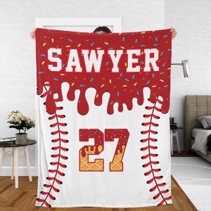 May include: A white baseball-themed blanket with red accents. The top features the name "SAWYER" in white block letters, with a red dripping design and colorful sprinkles. The number "27" is in a waffle-like font.