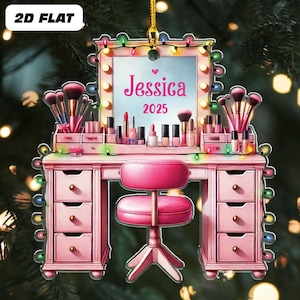 May include: A pink vanity ornament with a mirror, makeup, and a chair. The mirror has the name "Jessica" and the year "2025". The ornament is decorated with colorful lights and makeup brushes.