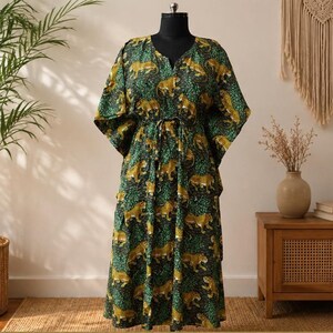 May include: A floor-length kaftan dress featuring a repeating pattern of yellow and brown leopards set against a dark green and black foliage background. The garment has wide, loose sleeves, a V-neckline, and a drawstring waist tie at the center.