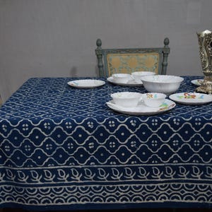 May include: A dining table set with a navy blue tablecloth featuring a white geometric pattern. White bowls, plates, and cups are arranged on the table. A decorative silver vase and a floral patterned chair are also visible.