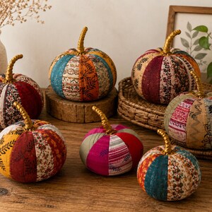 Puede incluir: Una colección de siete calabazas de tela con varios patrones de patchwork en tonos rojo, naranja, amarillo, azul y rosa. Cada calabaza tiene un tallo dorado texturizado y está construida con múltiples segmentos de tela verticales.