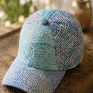 Puede incluir: Una gorra de béisbol con un diseño de patchwork de varios paneles de tela en tonos azul y verde azulado. La gorra está confeccionada con intrincadas costuras blancas en toda la superficie, creando una apariencia texturizada. Un pequeño botón de tela azul oscuro se encuentra en la corona.