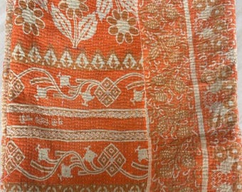 May include: A rectangular orange textile featuring a repeating pattern of white floral motifs, geometric shapes, and scrolling vines. The fabric displays visible horizontal running stitch lines throughout, creating a quilted texture with frayed edges.