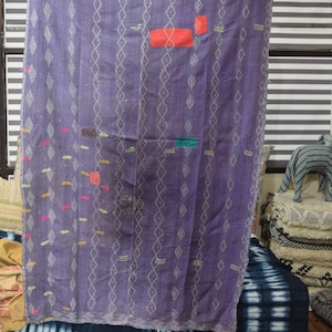 May include: A purple, handmade quilt with white diamond and vertical stitch patterns. The quilt features patches of red, green, and yellow fabric. The quilt is draped over a blue and white tie-dye fabric.