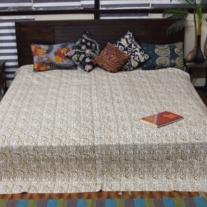 May include: A bed covered with a white and gold patterned quilt. Several decorative pillows with floral and geometric designs are arranged at the head of the bed. A book with a red cover rests on the quilt.