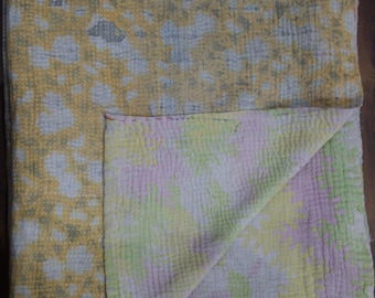 May include: A rectangular quilted blanket featuring a yellow and grey mottled pattern on the top side. The folded corner reveals a reverse side with a soft floral print in shades of pale pink, light green, and yellow, finished with visible stitching.