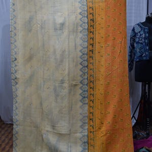 May include: A vintage, cream-colored textile with a blue patterned border and an orange patterned section. The textile has a textured appearance and appears to be handmade. The orange section has a repeating pattern.