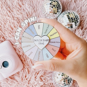 May include: A colorful wheel with the text "Taylor Swift Eras" is held in front of a pink fluffy background. The wheel is divided into sections with album names. The words "I CAN SPIN!" are also visible.