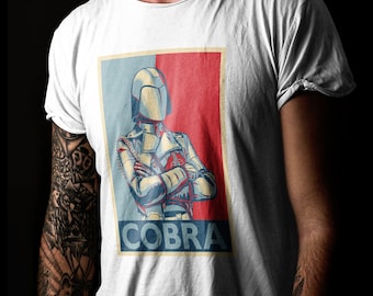 May include: A white t-shirt featuring a red, white and blue graphic of a person wearing a helmet with the word "COBRA" printed below.